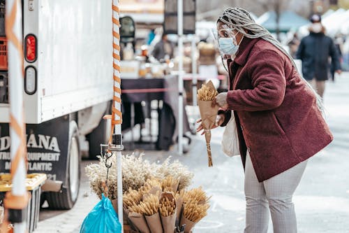 Black adult lady in warm coat and protective mask standing in street market and choosing bouquet of dried corn in daytime