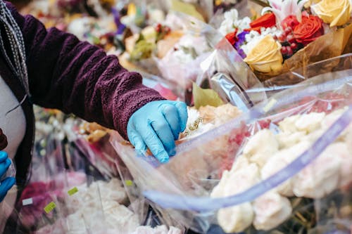 Anonymous female in casual clothes and medical gloves picking up flowers in floral shop in open market on street