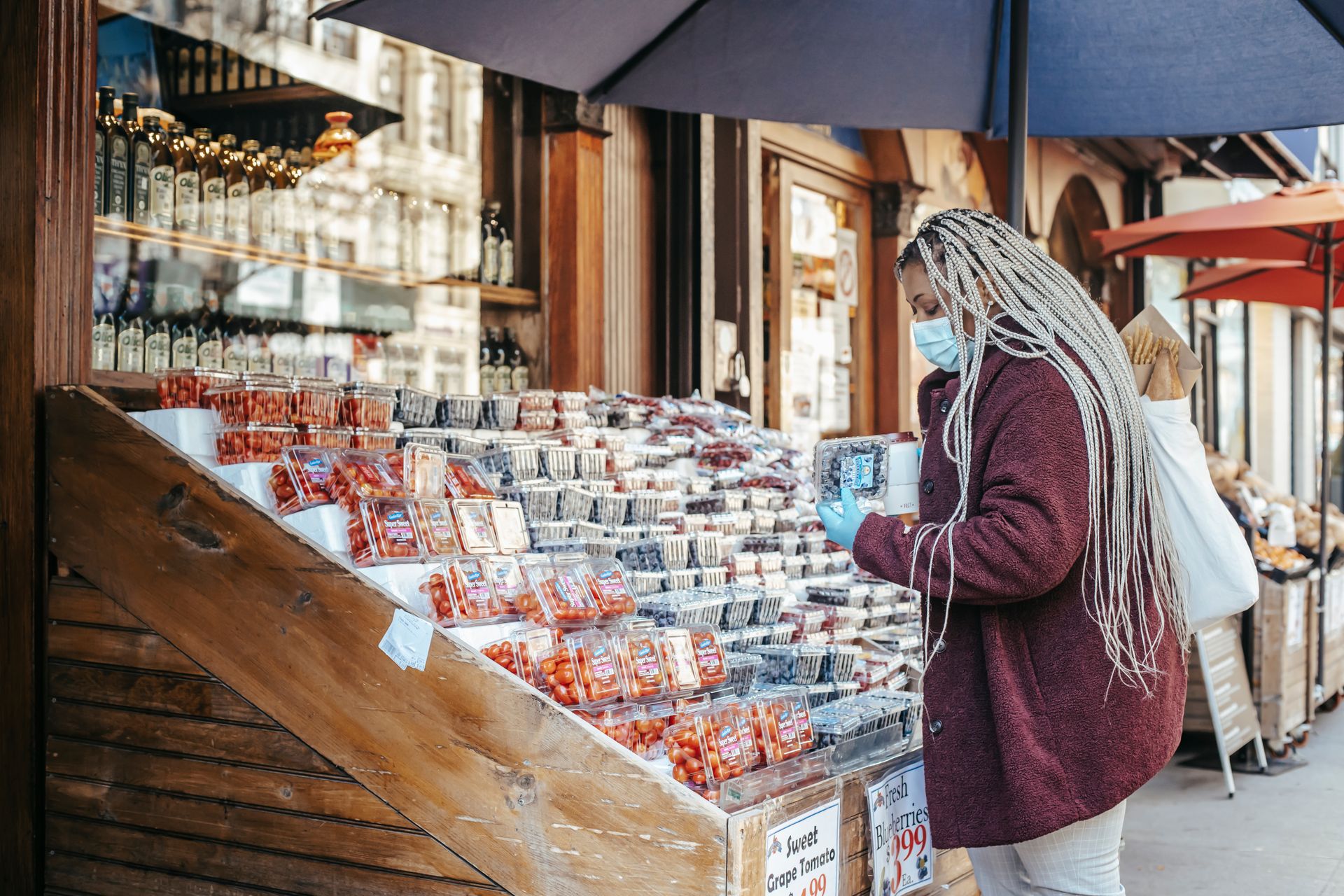 Side view of African American female in casual clothes mask and latex gloves standing near street stand with assorted food and choosing blueberries