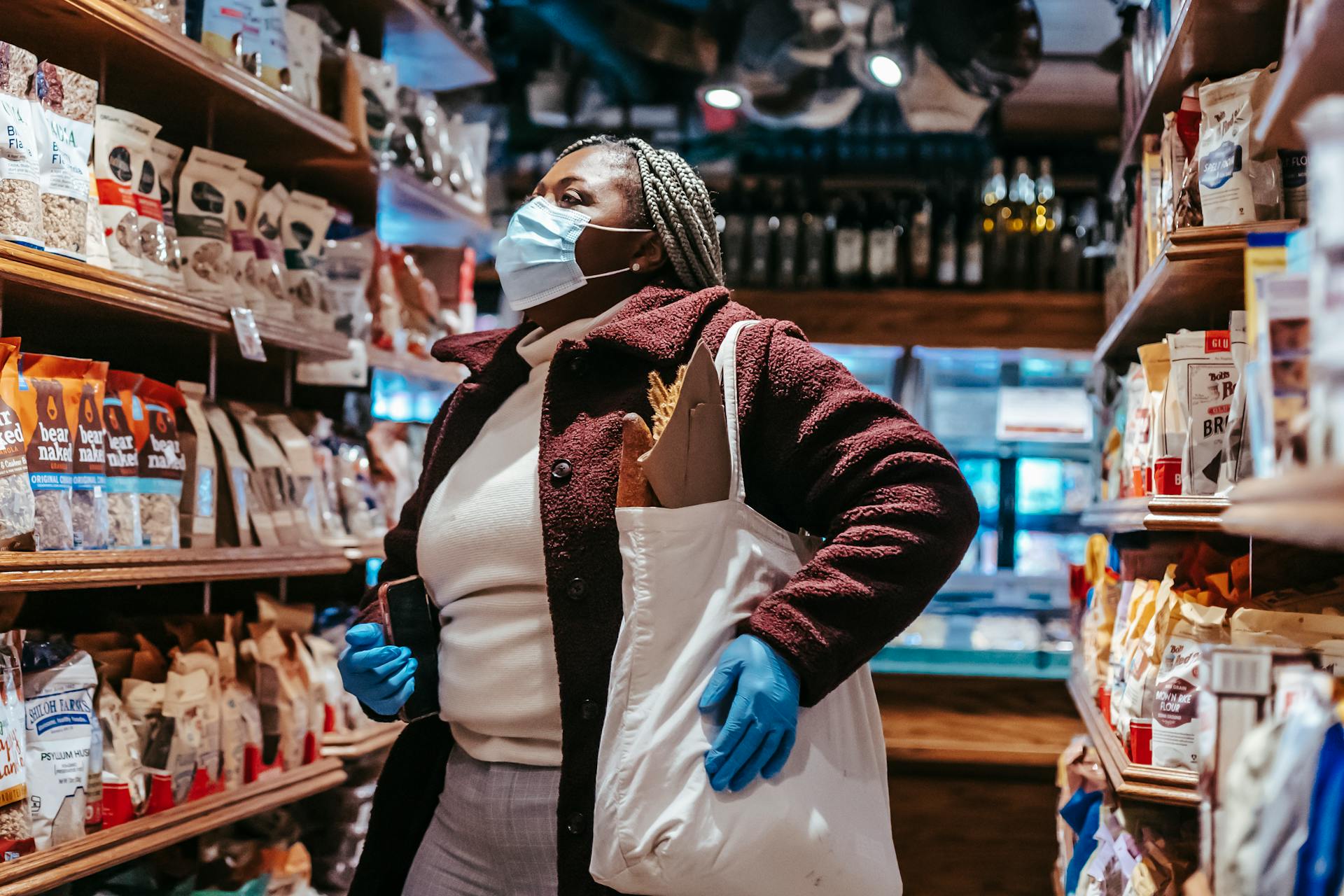 African American female buyer with shopping bag and smartphone in protective mask and gloves selecting products in supermarket