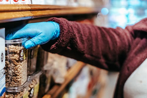 Crop anonymous woman in latex gloves picking food in plastic container while shopping in grocery store