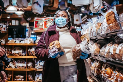 African American female buyer in protective mask and gloves carrying loafs of bread in grocery store