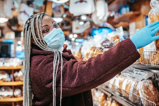 Side view of African American female buyer wearing medical mask and latex gloves choosing bread from shelf in supermarket