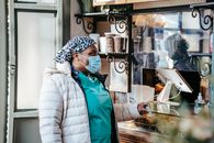 African American female medical worker in mask and uniform buying takeaway drink in coffee house