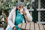 Adult African American lady in nurse uniform and jacket with mask and headband sitting at wooden round table in street cafe with cup of drink in daytime near green plants