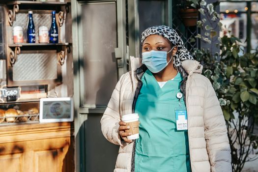 Adult black woman in nurse uniform with jacket and protective mask with headwear in city street with cup of drink near counter in sunny day