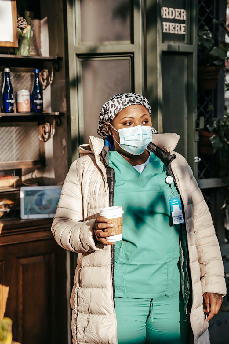 Black Nurse In Outerwear And Mask Standing In Modern Cafeteria