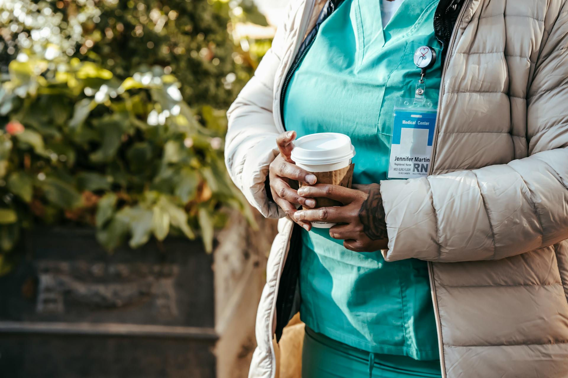 Crop anonymous African American nurse wearing warm clothes standing with coffee to go on street on sunny autumn day