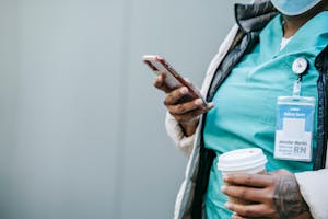 Crop faceless black nurse with takeaway coffee using smartphone