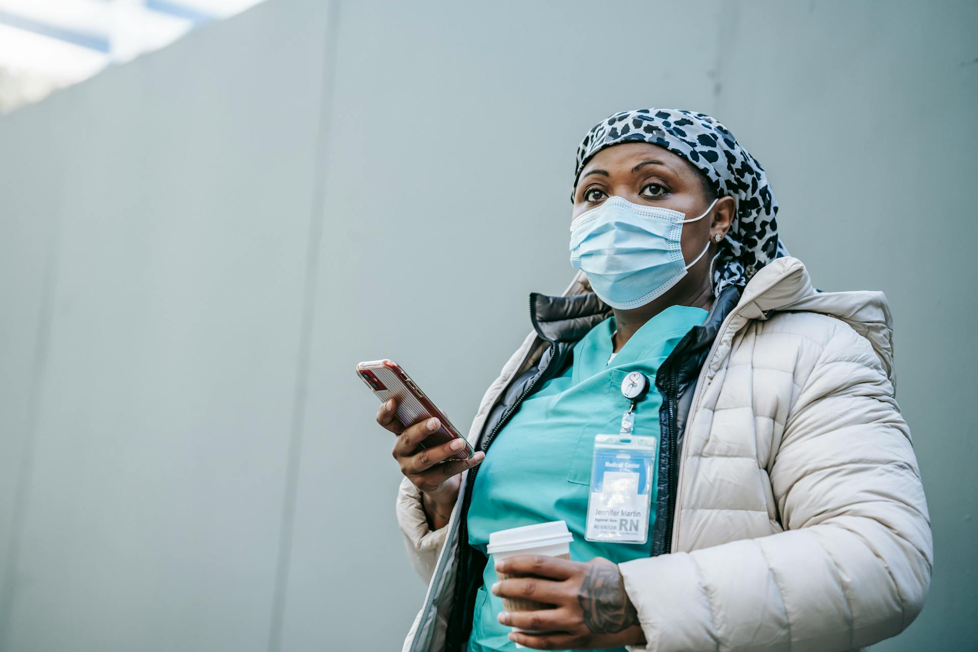 Serious adult African American nurse in warm clothes and protective face mask standing with cup of takeaway coffee and browsing mobile phone on street