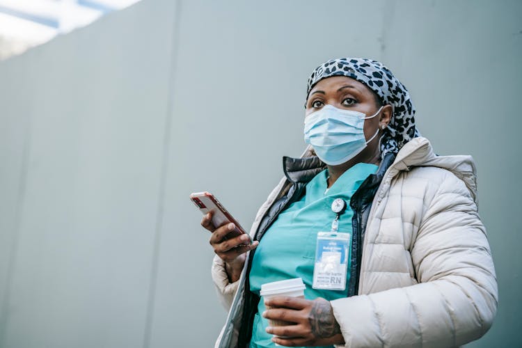 Black Nurse In Mask And Outerwear Using Smartphone