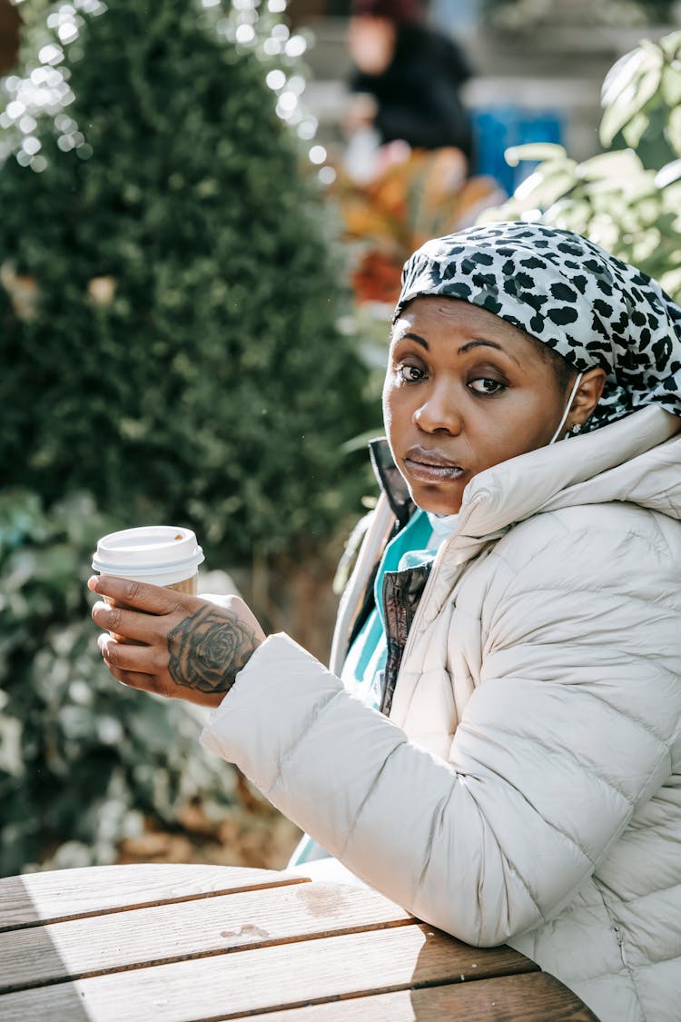 Contemplative Black Nurse In Outerwear Drinking Coffee In Outdoor Cafe