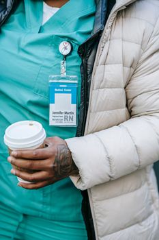 Anonymous African American nurse in blue uniform and warm wear standing with takeaway coffee on street