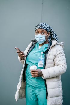 Adult African American nurse wearing uniform outerwear and mask standing with cup of takeaway coffee and browsing mobile phone on street