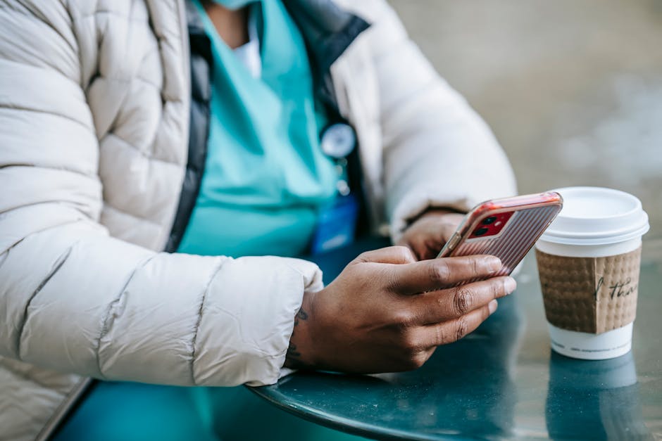 Crop anonymous African American female doctor browsing mobile phone while sitting at table with takeaway coffee in outdoor cafeteria