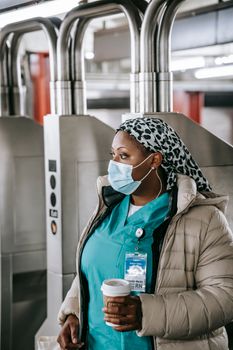Thoughtful adult African American female doctor in blue uniform warm wear and protective mask passing through turnstile in subway station and looking away