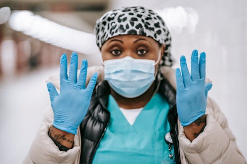 Pensive adult African American nurse wearing uniform warm clothes and face mask showing hands in blue latex gloves while standing in public hallway and looking at camera