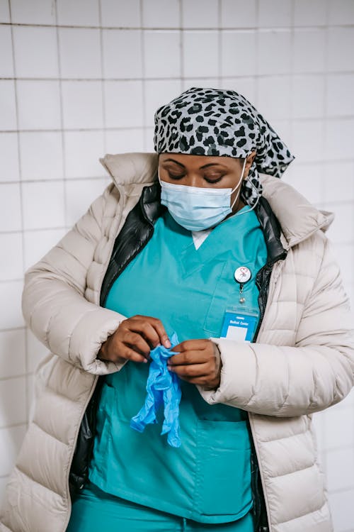 Adult plump African American nurse in warm clothes and protective mask putting on latex gloves while standing in hallway in public place