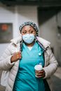 Contemplative adult African American nurse with takeaway coffee wearing medical mask and warm clothes standing in hallway in public place