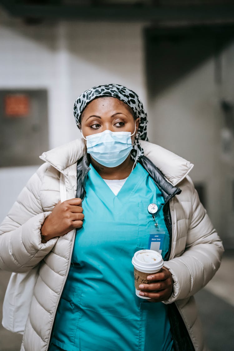 Calm Black Nurse In Outerwear And Mask Standing In Hallway