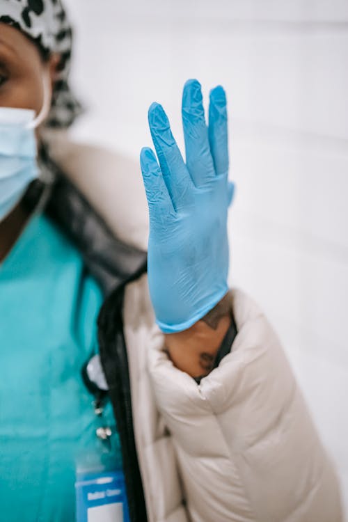 Crop African American nurse in outerwear and face mask raising hand in blue latex glove while standing against white wall in public place
