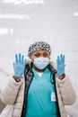 Calm adult African American nurse in uniform warm clothes and face mask raising arms and showing hands in latex gloves while standing in public place and looking at camera