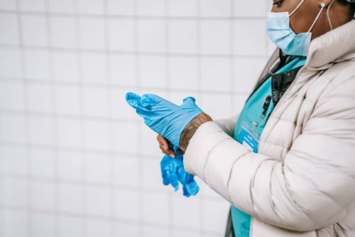 Free Side view crop African American nurse in warm clothes and protective mask putting on latex gloves while standing in public place corridor Stock Photo