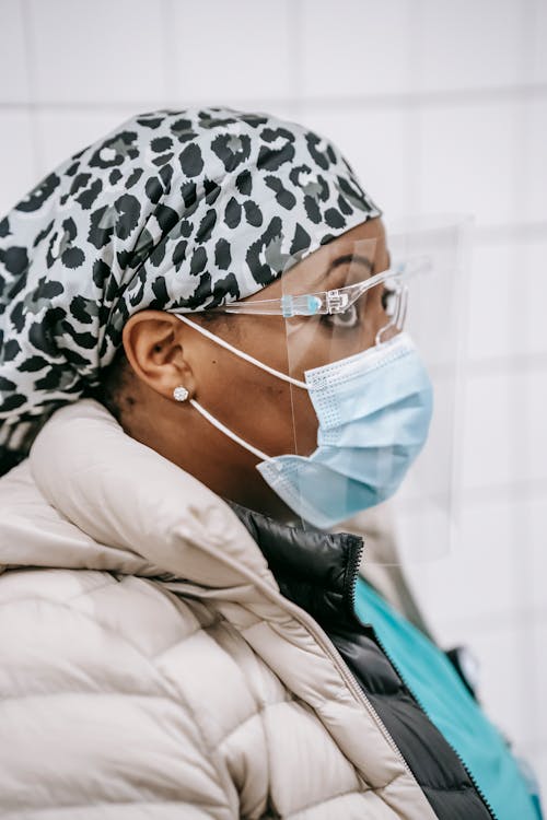 Side view of black woman in medical mask and protective glasses standing in tiled room and looking away