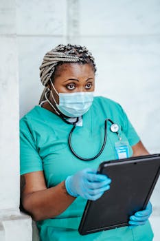 Female nurse in scrubs and mask reviewing patient data on a tablet.