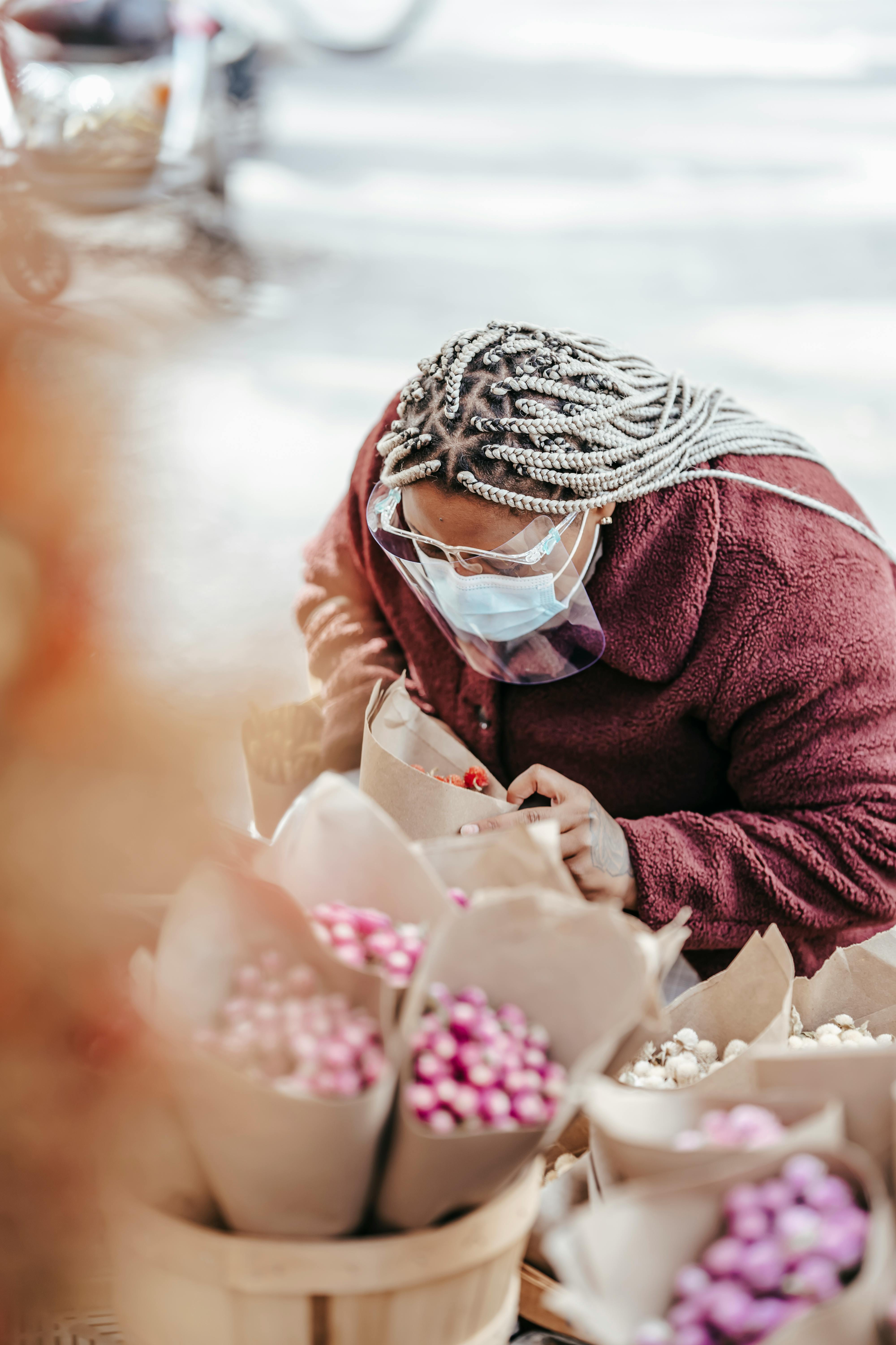 Unrecognizable ethnic woman with braids in protective mask standing on street and choosing bouquet of flowers in floristry store in daytime