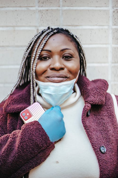 Cheerful African American woman with braids in protective mask and gloves standing with smartphone against brick wall and looking at camera