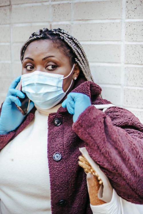 Free Pensive African American woman in medical mask and casual clothes standing against brick wall and talking on mobile phone Stock Photo