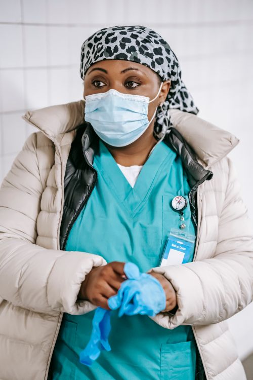 Free Pensive black woman doctor in protective mask and uniform standing and putting on medical gloves against light wall Stock Photo
