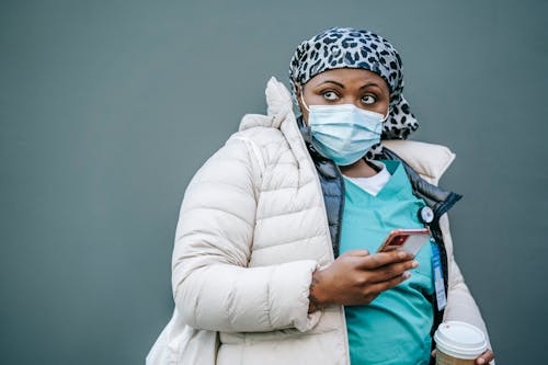 Free Pensive African American woman doctor in medical mask and uniform standing with cup of takeaway coffee and smartphone and looking away Stock Photo