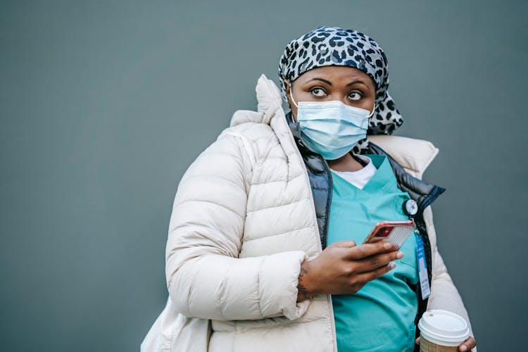 Black Woman Doctor In Mask Standing With Cup Of Coffee And Smartphone