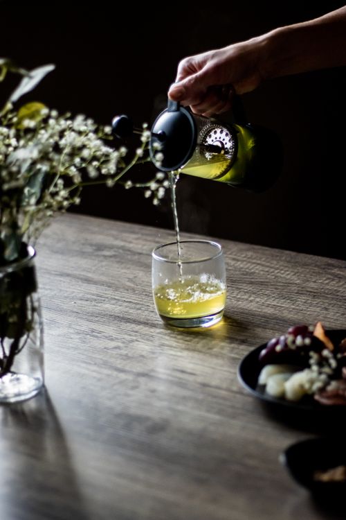 Free A close-up shot of herbal green tea being poured into a drinking glass on a wooden table, with flowers and snacks. Stock Photo