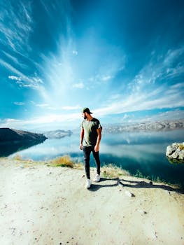 A man in casual attire stands on a cliff with a breathtaking view of a clear lake under a vibrant blue sky.