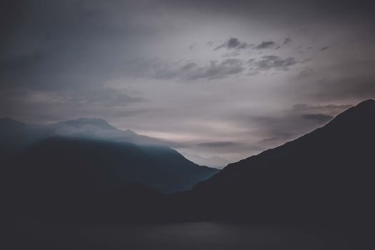 Dramatic view of dark mountain range with peaks covered with mist against gloomy cloudy sky in evening