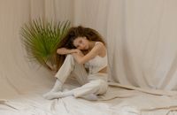 Woman in White Tank Top and White Pants Sitting on White Sheets