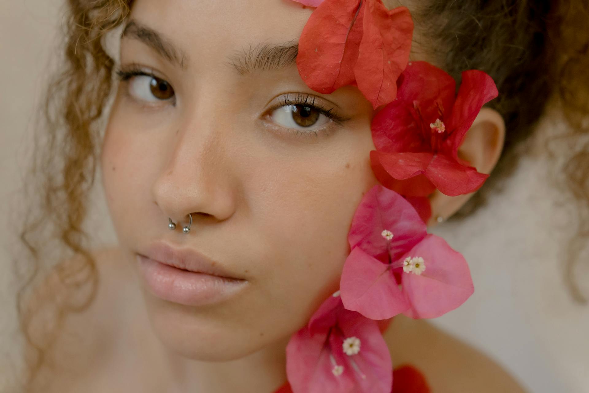 A woman with curly hair and septum piercing adorned with vibrant red bougainvillea flowers.