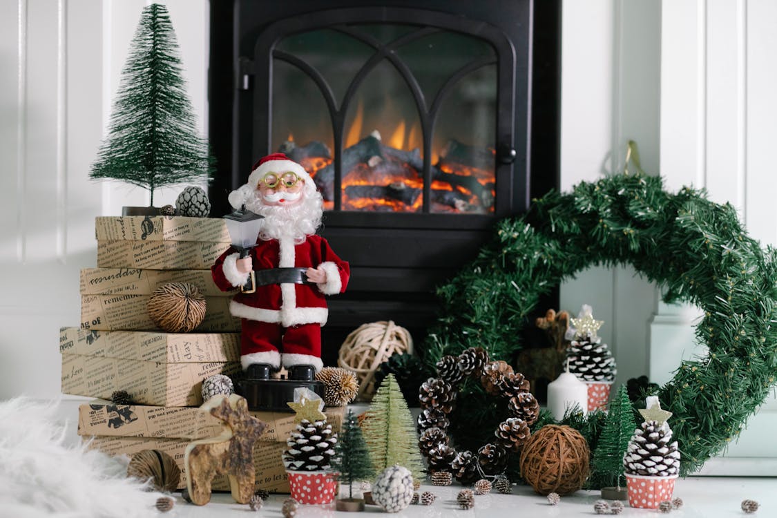 Statuette of Santa Claus placed on wrapped gift boxes near Christmas decorations at warm fireplace with green wreath at home