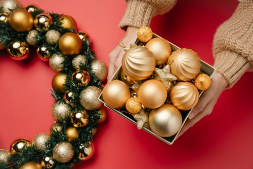 Free Top view of anonymous female with box of golden baubles and stars in hands on red background with green wreath decorated with balls Stock Photo