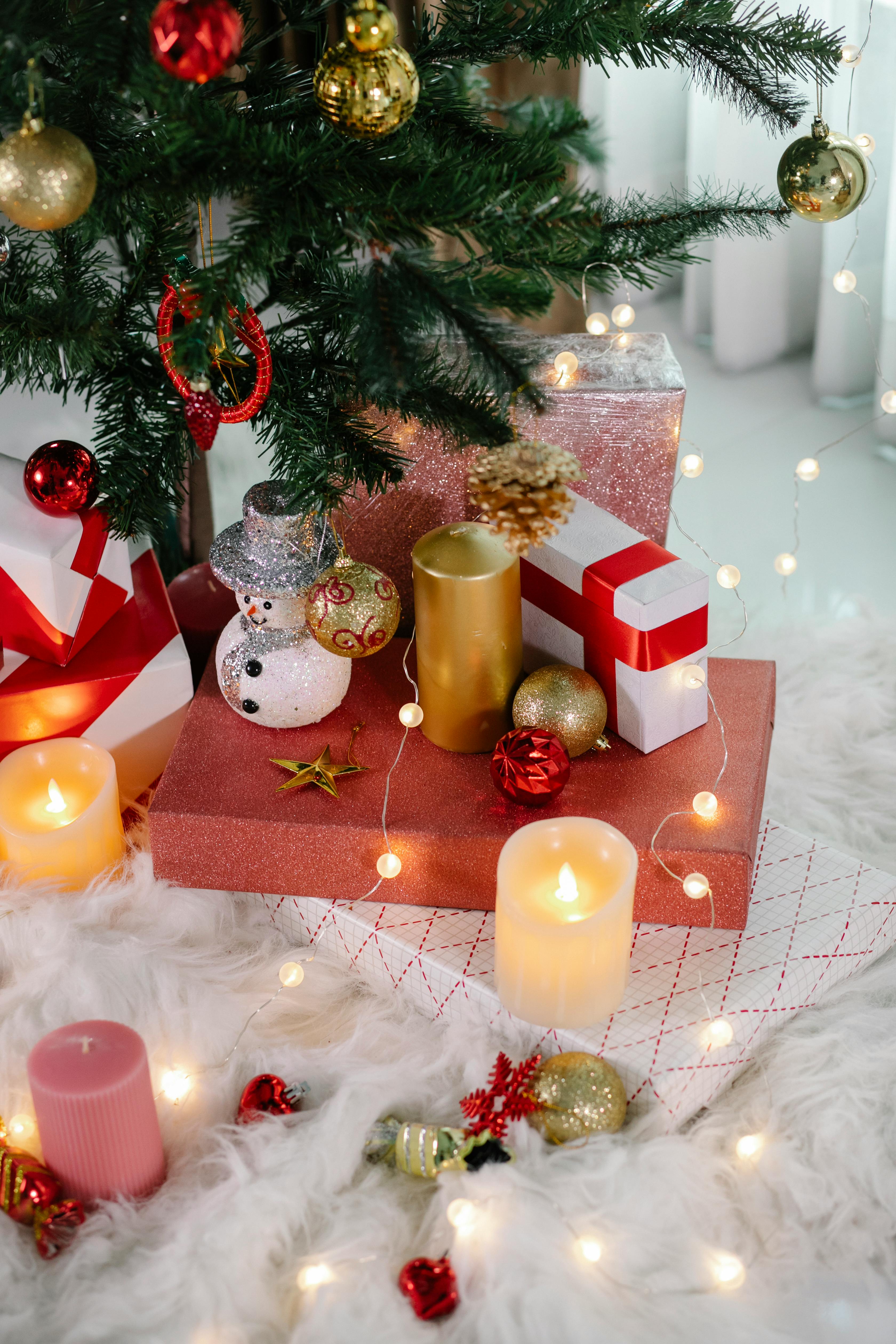 From above of gift boxes and decorations placed on white rug under Christmas tree with baubles in cozy room with burning candles and glowing garland
