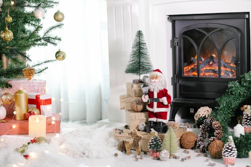 Cozy light room with gift boxes under Christmas tree and Santa Claus near holiday decorations placed on white cozy fabric at home with fireplace and burning candles