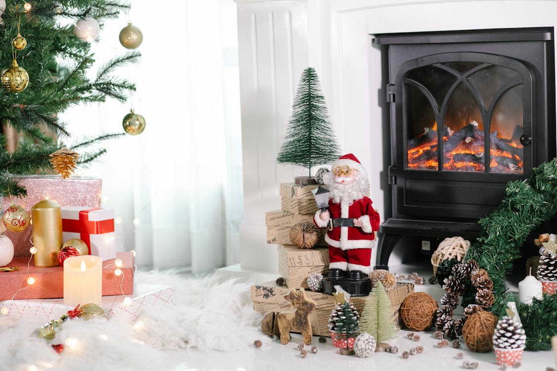 Free Cozy light room with gift boxes under Christmas tree and Santa Claus near holiday decorations placed on white cozy fabric at home with fireplace and burning candles Stock Photo