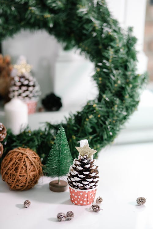 Small pine cone decorated with snow placed on floor near bauble and green wreath against Christmas decorations on blurred background