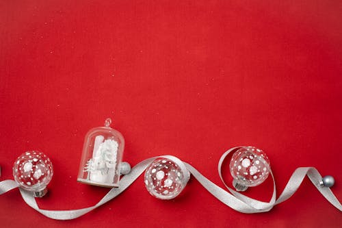 Top view of Christmas composition with small Christmas tree baubles and souvenir covered with glass dome and wavy silver ribbon on red background