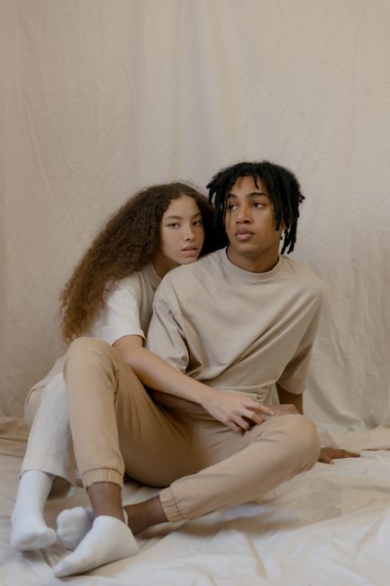 Interracial couple with natural hair sitting close on a beige background, showcasing intimacy and fashion.