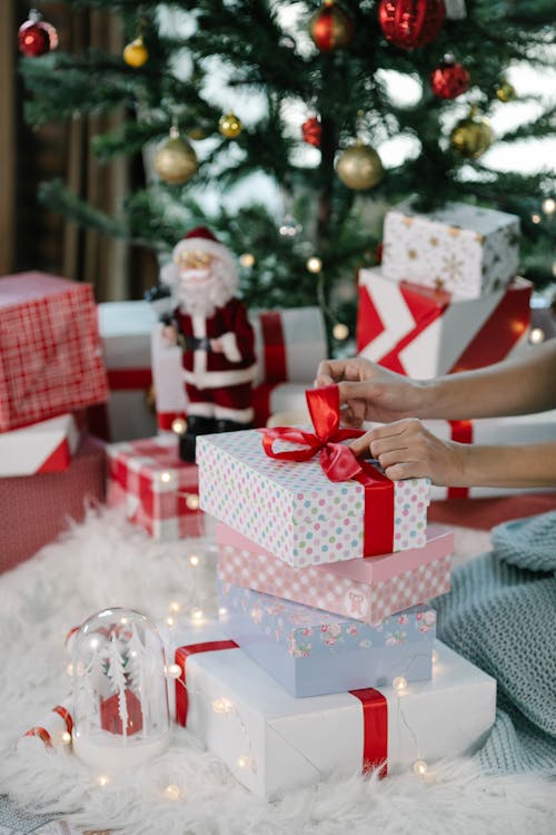 Crop anonymous female sitting near Christmas tree and tying red ribbon in bow on present box