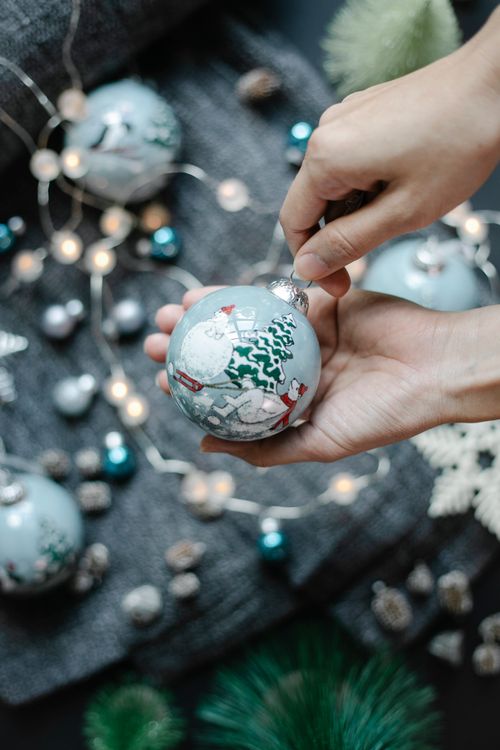 Free From above crop unrecognizable person with Christmas painted ball with polar bear near Christmas tree with snowman near different decorations and garland on gray textile Stock Photo